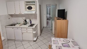 Een compacte keuken met witte kastjes, een magnetron en een koffieapparaat. Er staat een klein tafeltje met een tafelkleed met dessin en een televisie op een houten dressoir.