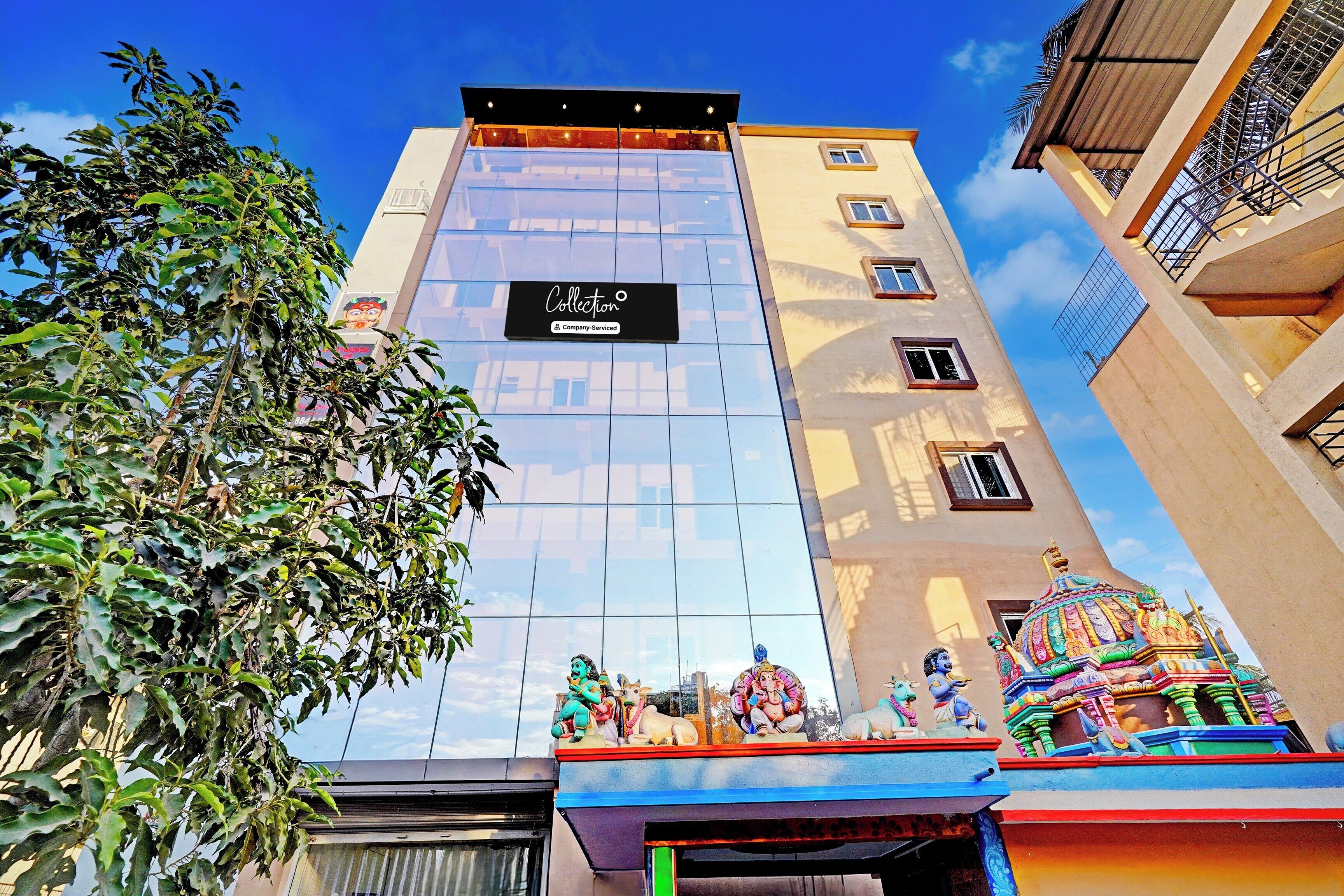 A hotel with a modern facade, a sign indicating it is a company-serviced collection, and a colorful statue of deities on the ground level.