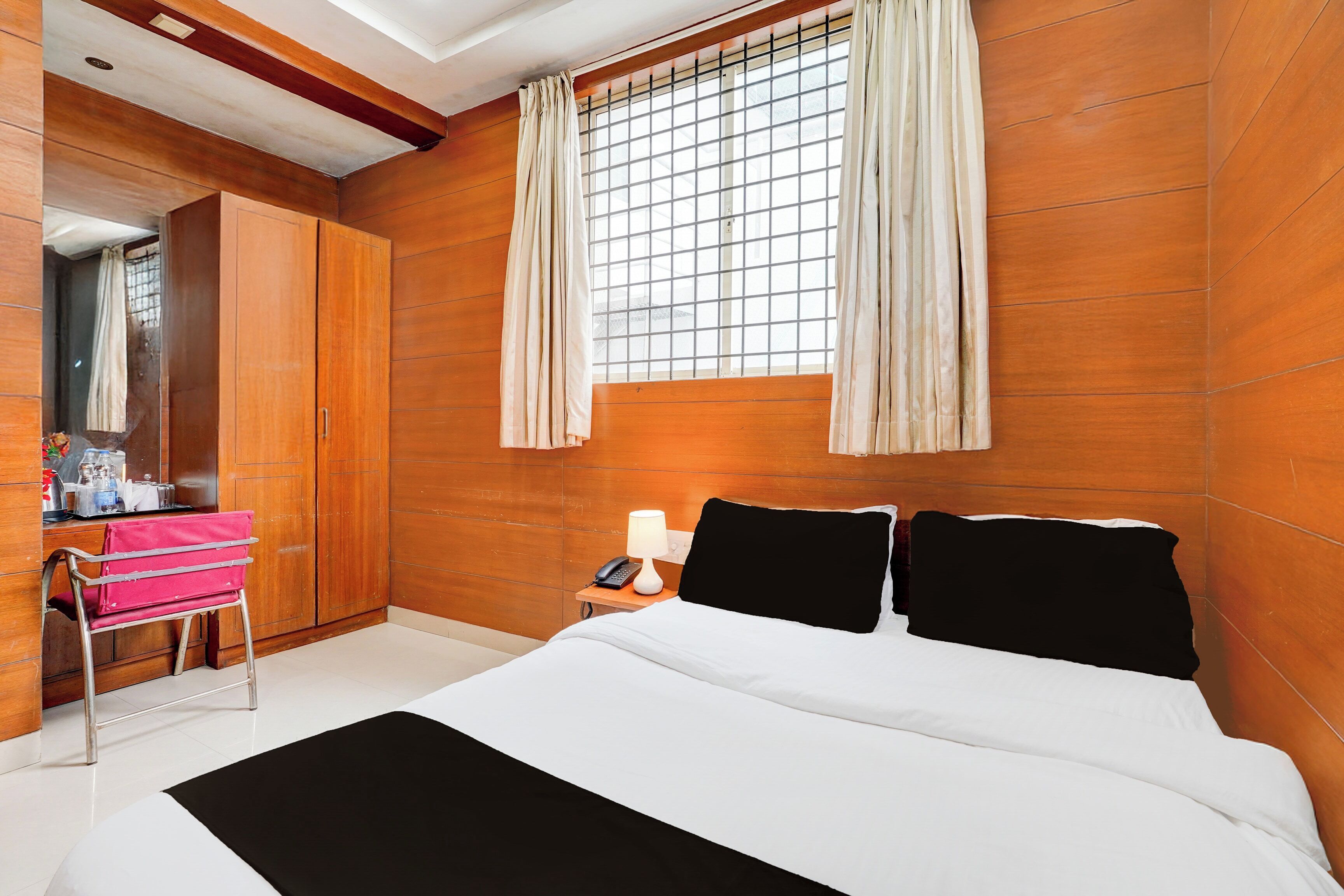 A hotel room with a bed, a bedside table with a lamp, a mirror, a wardrobe, and a window with curtains.