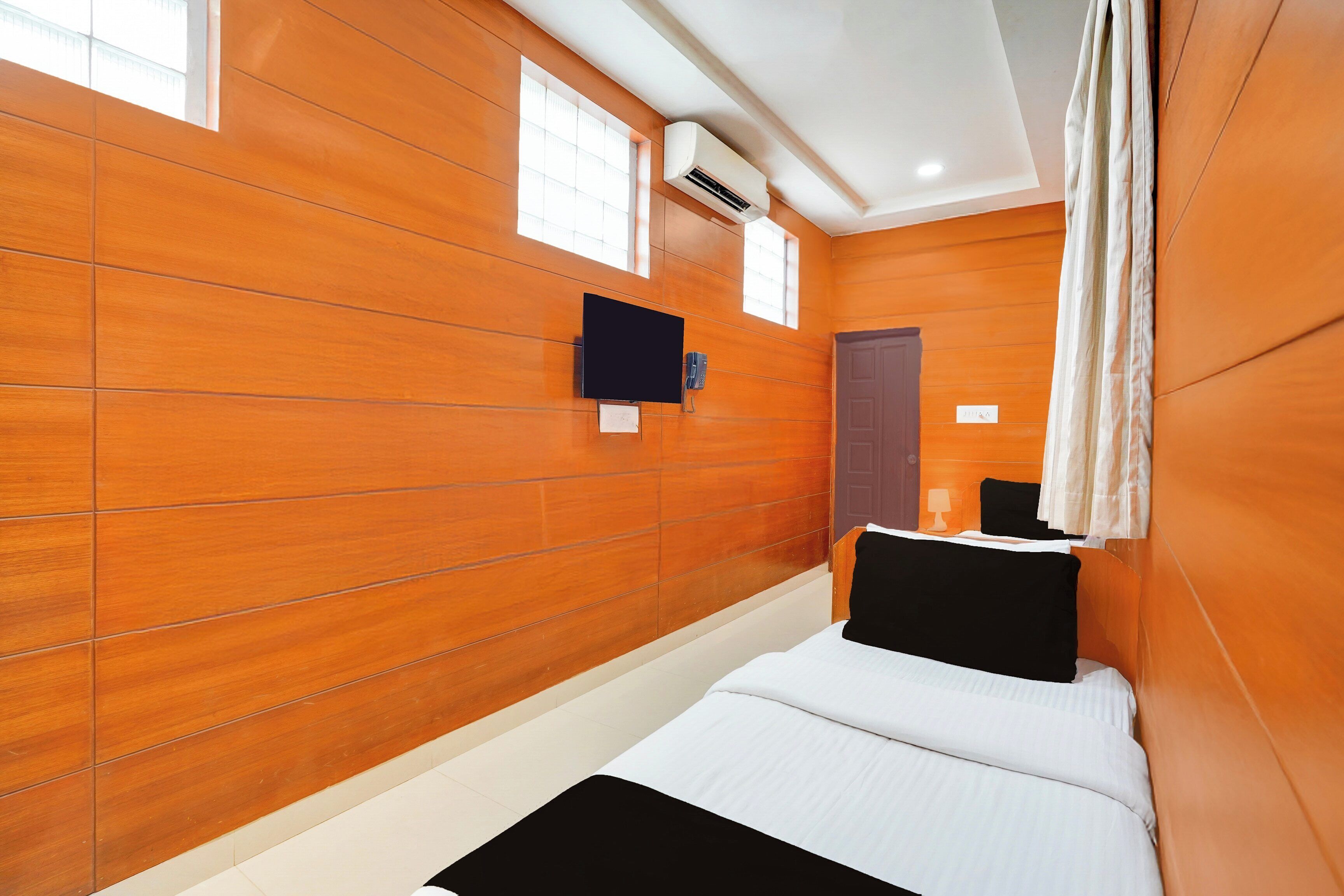 A hotel room with a wooden wall, a bed with white and black bedding, a flat-screen TV mounted on the wall, and a window with curtains.