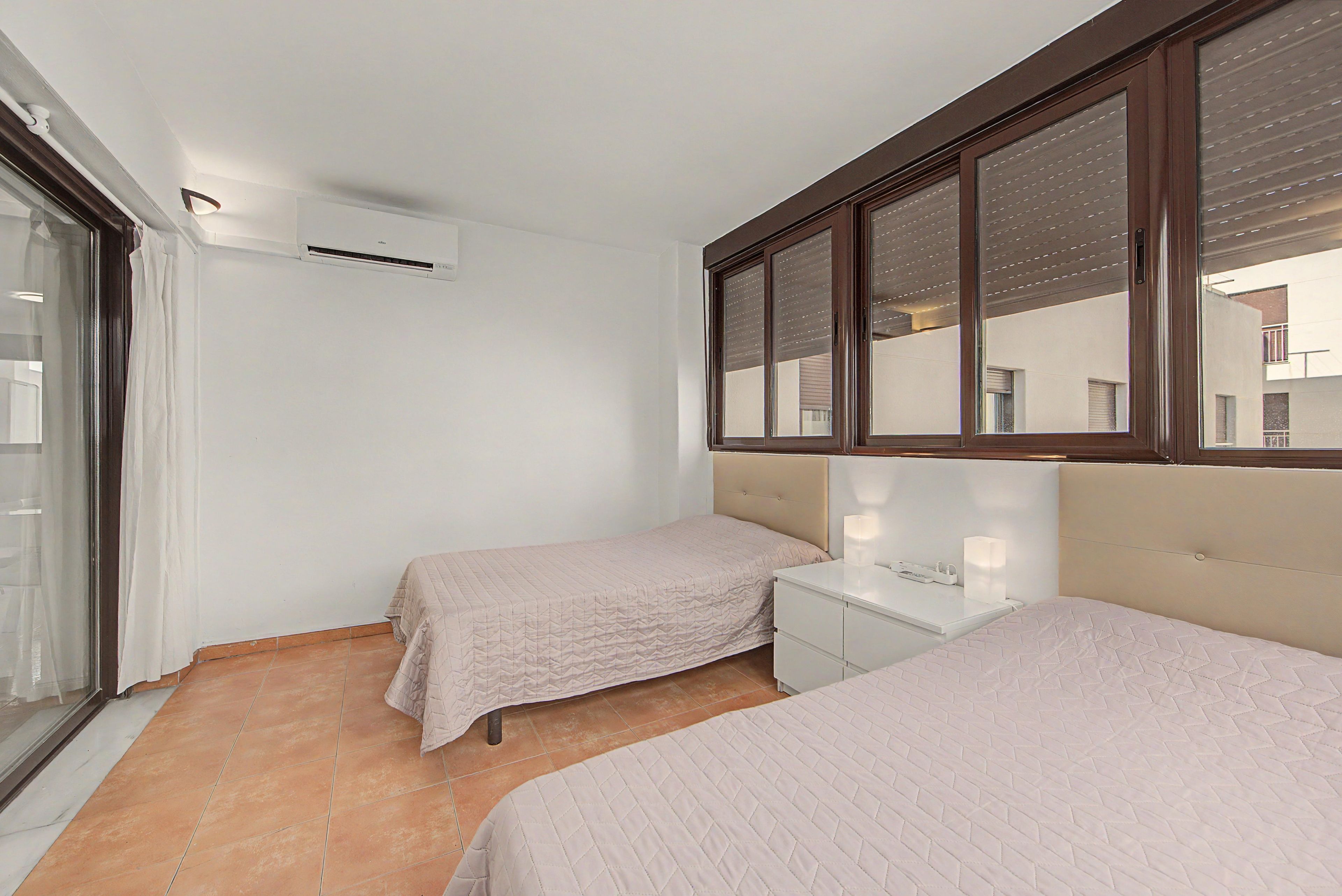 Une chambre avec deux lits, une table de chevet, une fenêtre avec des stores et un appareil de climatisation.