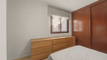 A bedroom with a wooden dresser, a bed with a patterned bedspread, a window with a blind, and a wooden wardrobe.