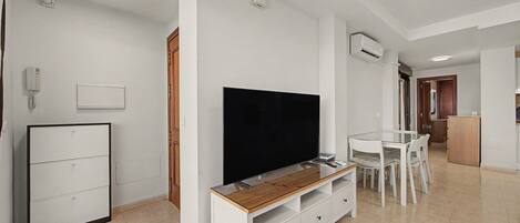 Una sala de estar moderna con un televisor de pantalla plana, un mueble de madera para entretenimiento, una zona de comedor con una mesa y sillas blancas, y una cocina con un armario de madera.