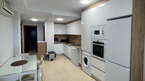A modern kitchen with white cabinetry, a built-in oven, microwave, and dishwasher. There is a dining area with a glass table and white chairs.