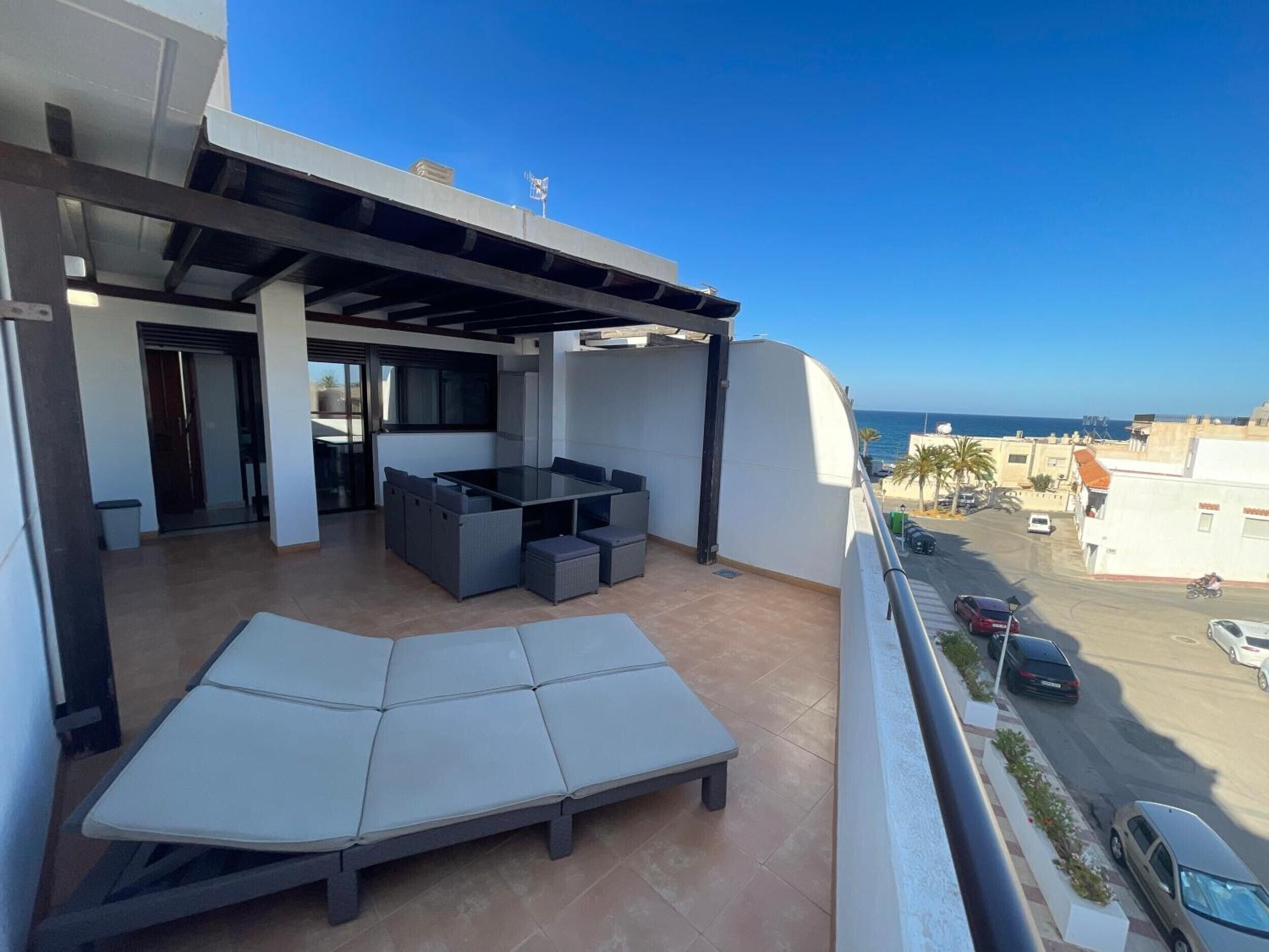 Een balkon met een bank en een tafel, met uitzicht op een straat en de zee.