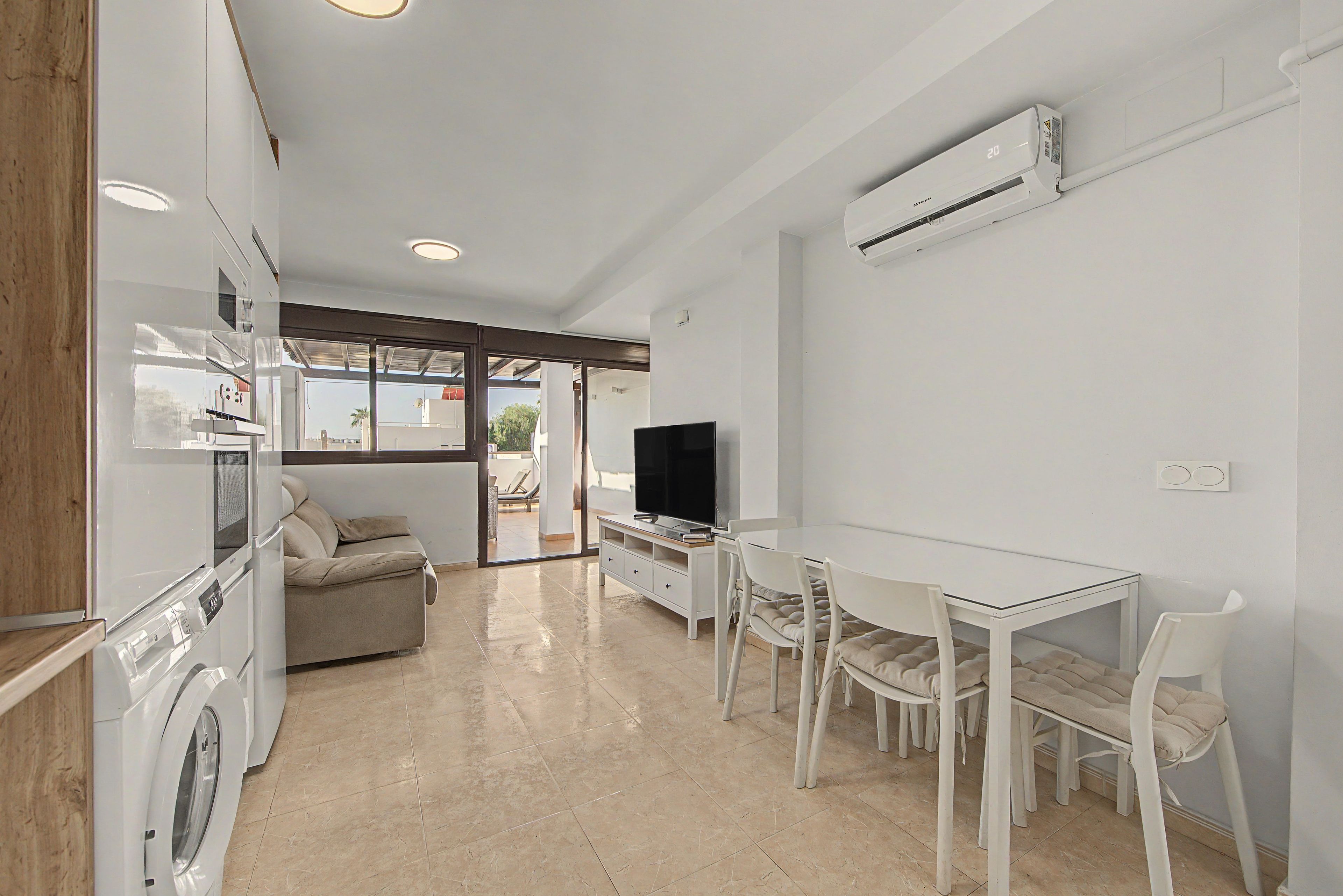 A modern kitchen with a built-in washing machine, a dining area with a glass table, and a flat-screen TV.