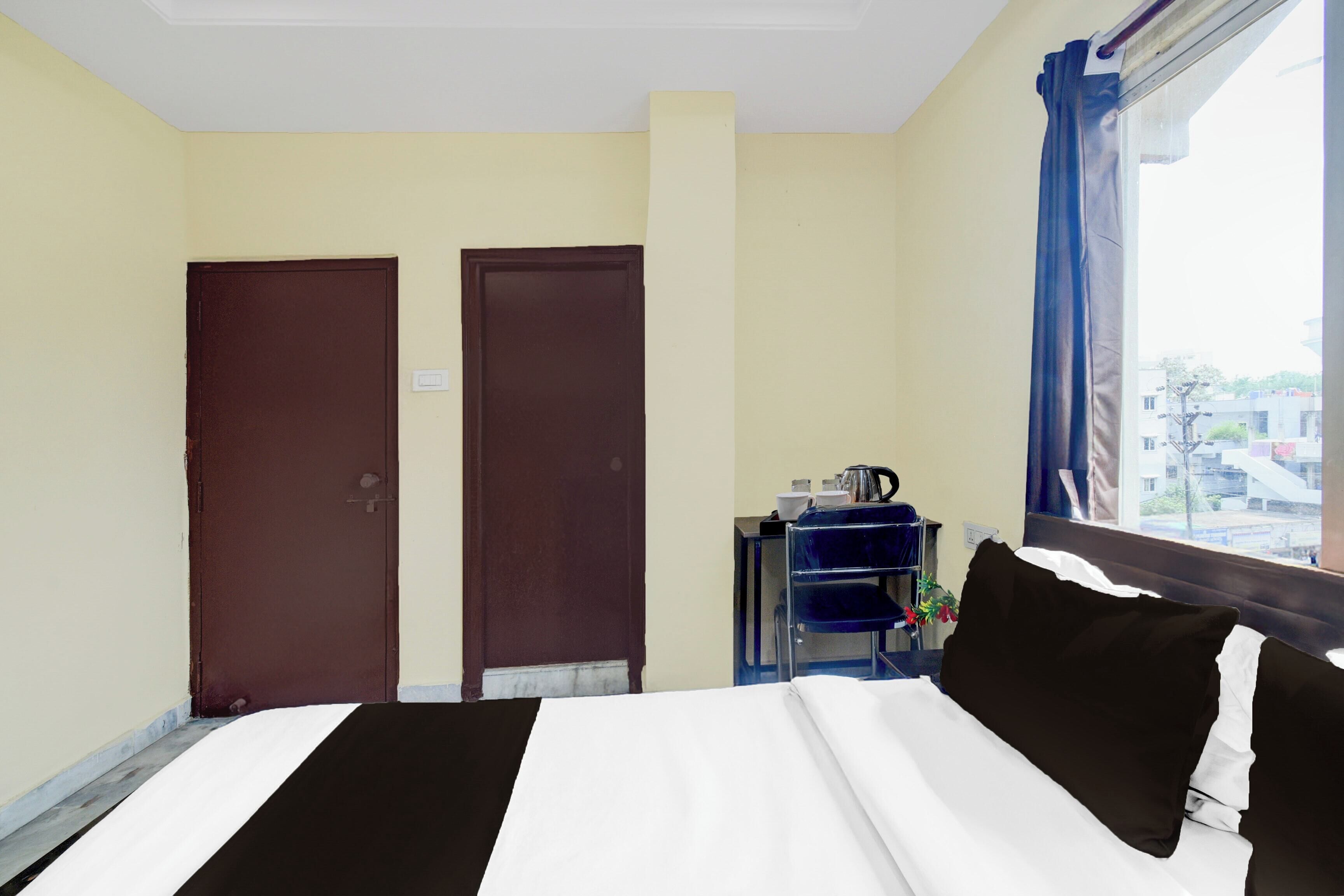 A hotel room with a bed, two doors, a window with curtains, and a small table with a kettle.