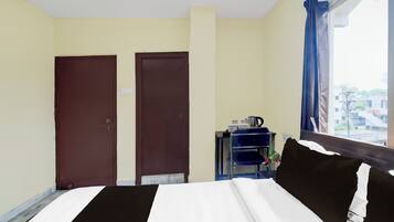 A hotel room with a bed, two doors, a window with curtains, and a small table with a kettle.