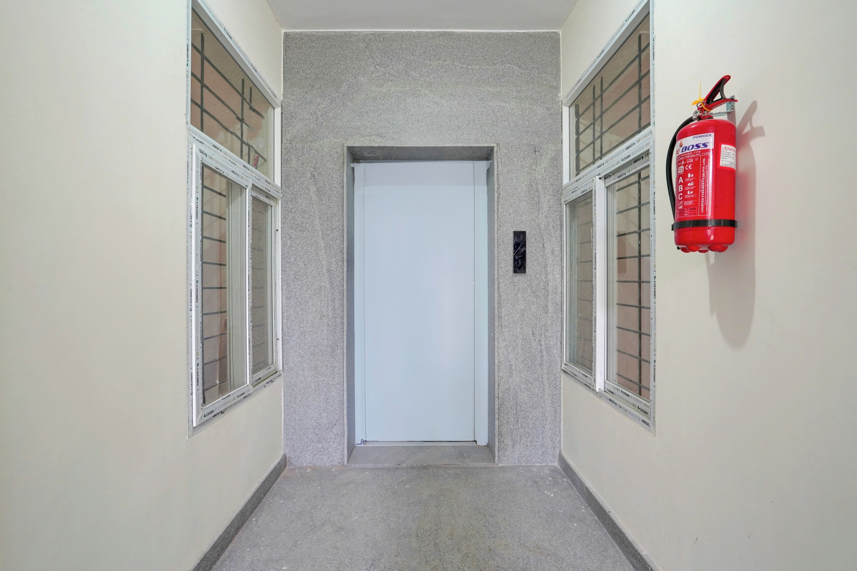 Un pasillo con un extintor montado en la pared, una puerta al fondo y ventanas con persianas a ambos lados.