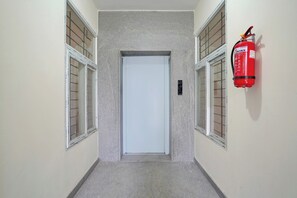 A hallway with a fire extinguisher mounted on the wall, a door at the end, and windows with blinds on either side. - Collection O REVA University Rukmini (Bangalore)