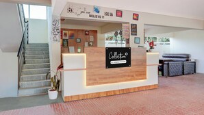 Un mostrador de recepción con un letrero que indica 'Collection' y 'Company-Serviced'. Hay carteles motivacionales en la pared, una escalera a la izquierda y una planta en maceta frente al mostrador.