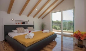 Ein Schlafzimmer mit einem großen Bett, Holzbalken, einer Schiebetür aus Glas und Blick ins Grüne.
