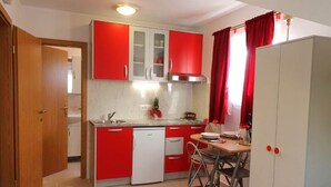 A compact kitchen with red cabinets, a white dishwasher, and a dining area with a table and chairs. - Apartments Clementina Sunrise-standard Studio Apartment (Orebic)