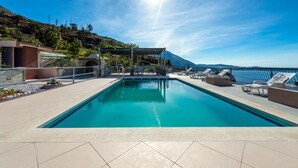 A rectangular swimming pool with a clear blue water surface, surrounded by a tiled deck. There are lounge chairs and a covered seating area with a view of the landscape. - Apartments Klis -two-bedroom Apartment With Terrace (Zupa dubrovacka)