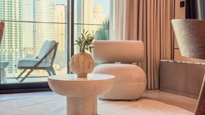 A modern living room with a view of a cityscape, a round table with a plant, a lounge chair, and a large ottoman. - Marina Living Residences - Dubai Marina (Dubai)
