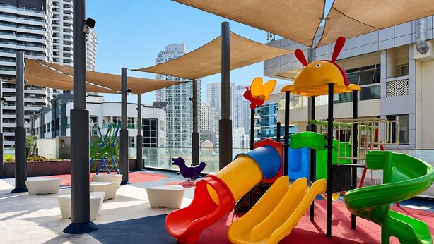 An outdoor play area with slides and climbing structures, surrounded by modern buildings. - Marina Living Residences - Dubai Marina (Dubai)