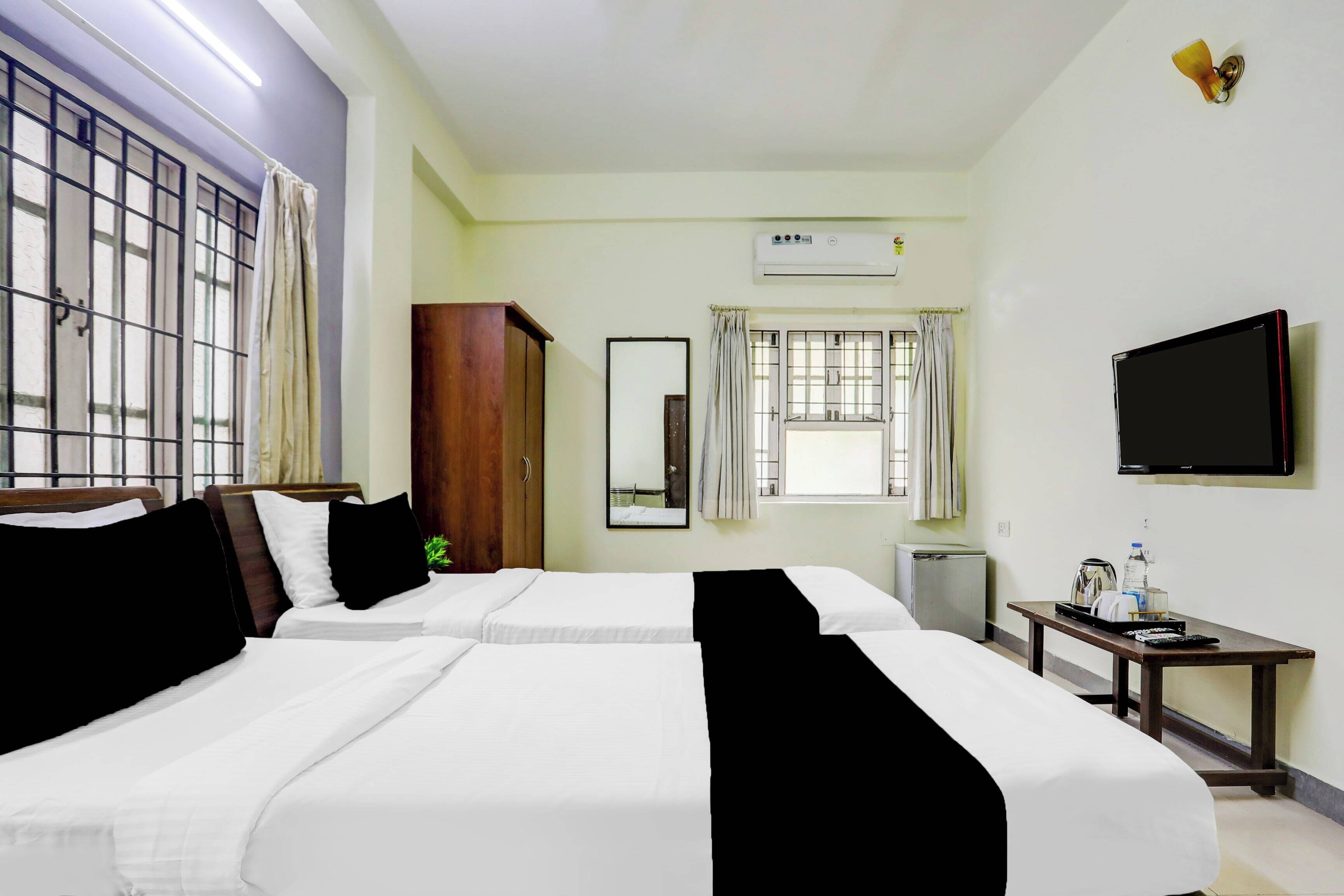 Habitación de hotel con dos camas, televisor, equipo de aire acondicionado, ventana con cortinas y una mesita con hervidor y botella de agua.