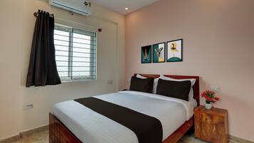 Una habitación de hotel con cama, una ventana con cortinas, un aire acondicionado de pared y una mesita de noche con un jarrón de flores.