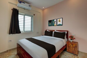 Quarto de hotel com cama, janela com cortinas, ar condicionado fixado na parede e mesa de cabeceira com um vaso de flores.