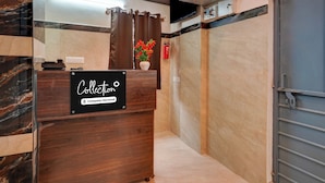Zona de recepción con mostrador de madera, un cartel que indica 'Compañía de Cobranza - Servicio', un jarrón con flores, un extintor y una puerta con picaporte metálico.