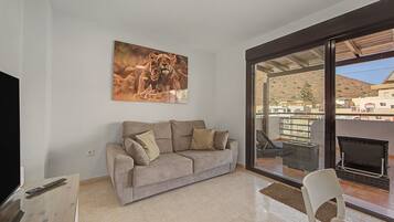 Een woonkamer met een beige bank, een televisie en een groot raam met uitzicht op een woonwijk.