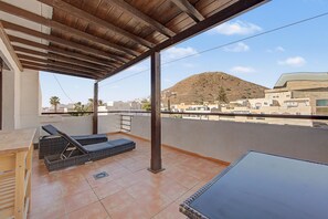 Un espace extérieur abrité, doté d’une pergola en bois, de deux fauteuils, d’une table et offrant une vue sur une colline parsemée de bâtiments.