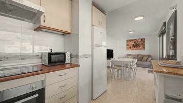A modern kitchen with white cabinets, a built-in microwave, and a refrigerator. There is a dining area with a table and chairs, and a sofa in the background.