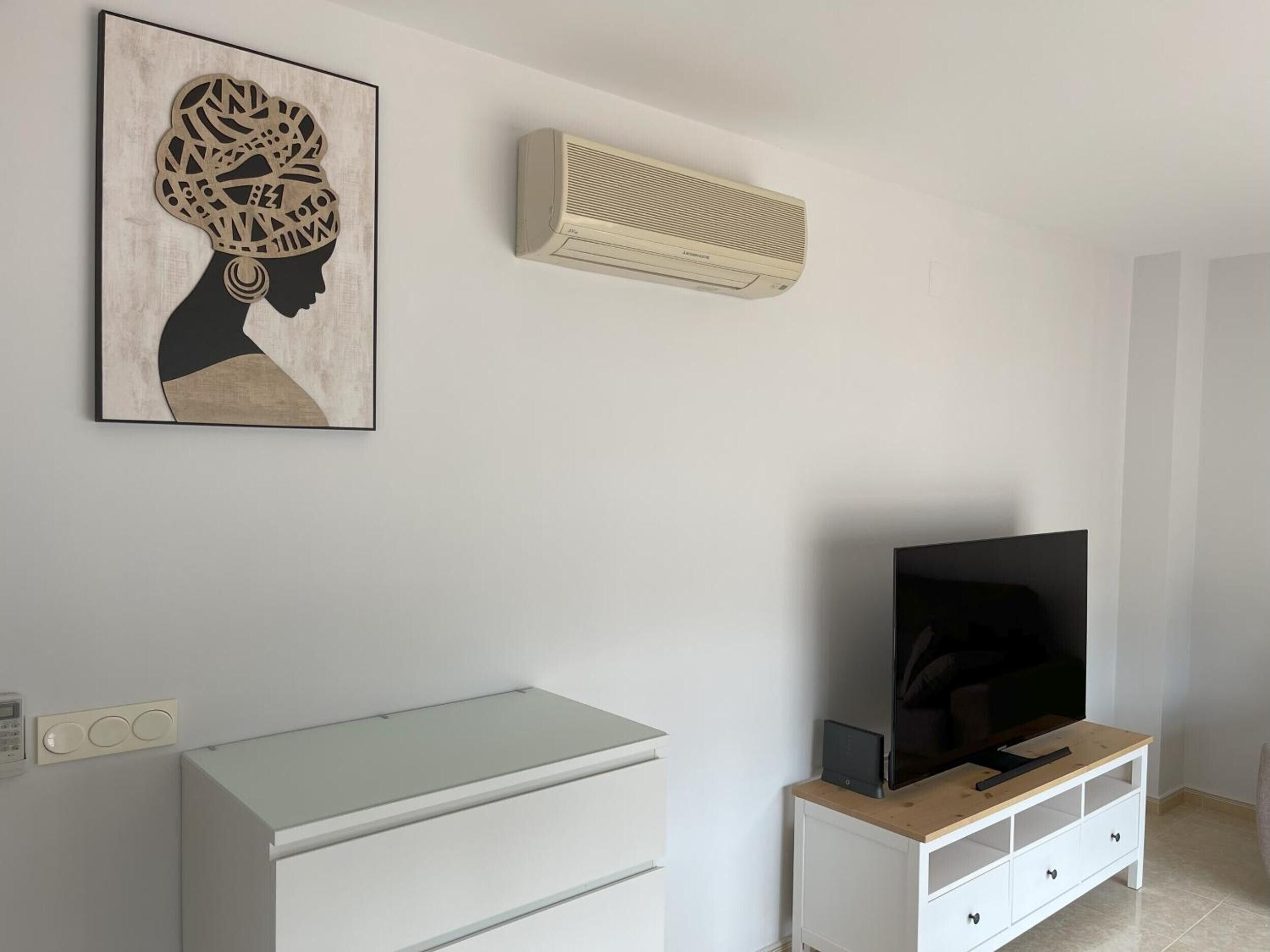 Ein modernes Wohnzimmer mit Flachbildfernseher auf einem Holz-TV-Möbel, einer Wand-Klimaanlage und einem gerahmten Kunstwerk über dem Fernsehmöbel.