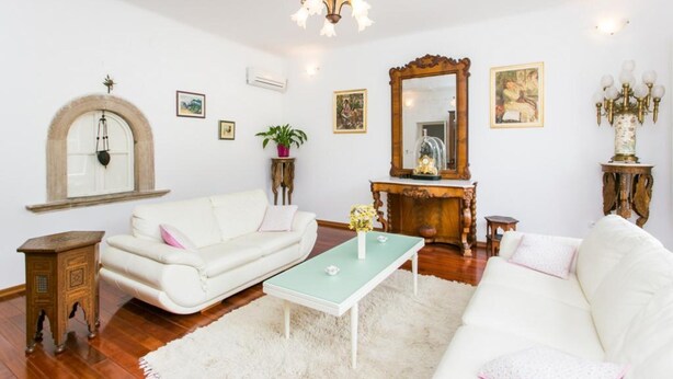 A living room with a white sofa, a green coffee table, a white rug, a wooden side table, a mirror, and a chandelier. - Villa Marini Dvori - Two-bedroom Apartment With Terrace (Dubrovnik)