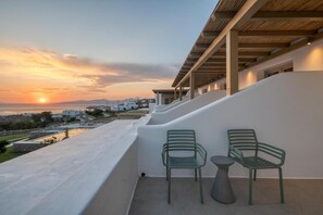 A balcony with two chairs and a table, overlooking a coastal area with buildings and a sunset. - Ostria View Resort (Mykonos)