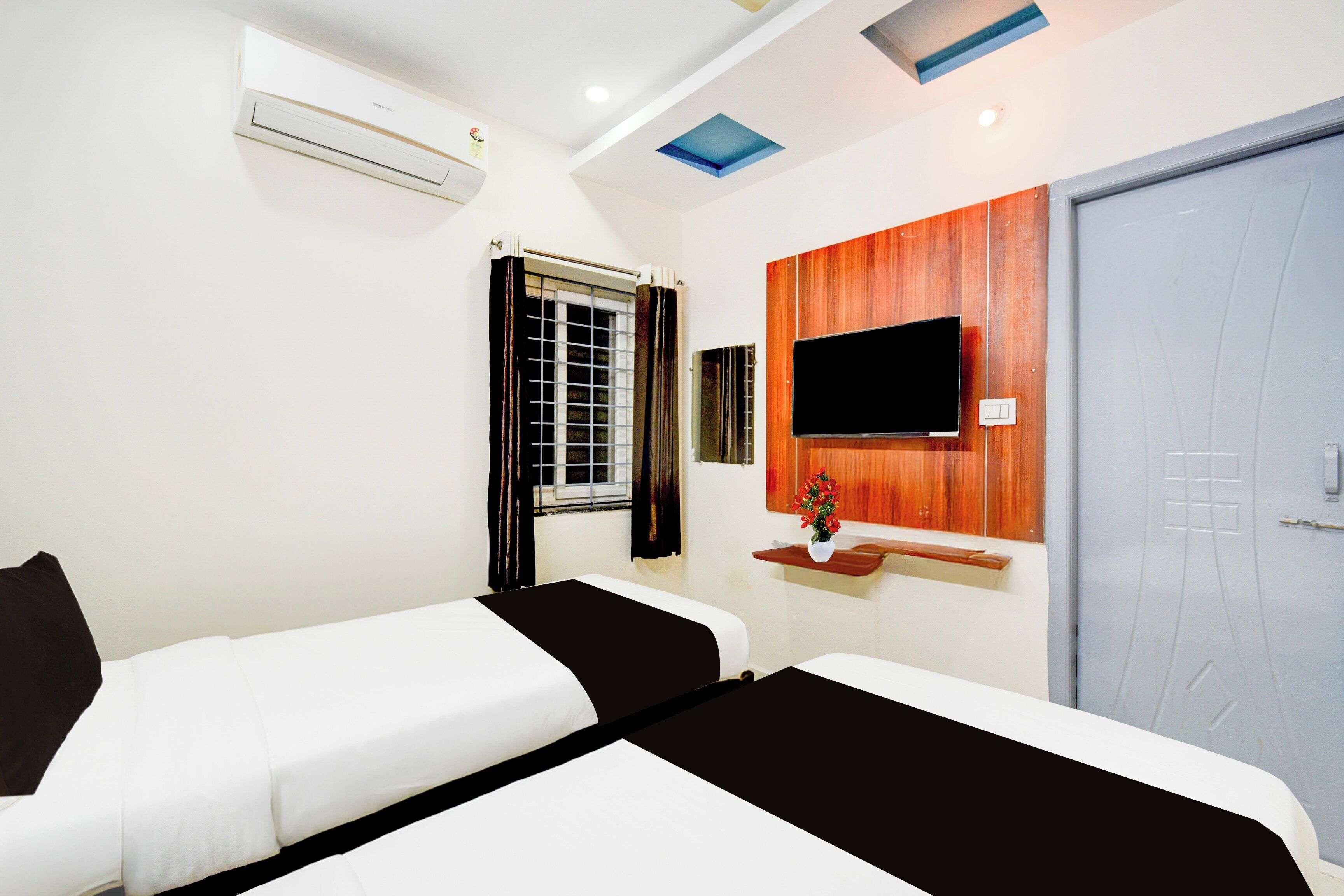 A hotel room with two beds, a TV mounted on the wall, a window with curtains, and an air conditioning unit.