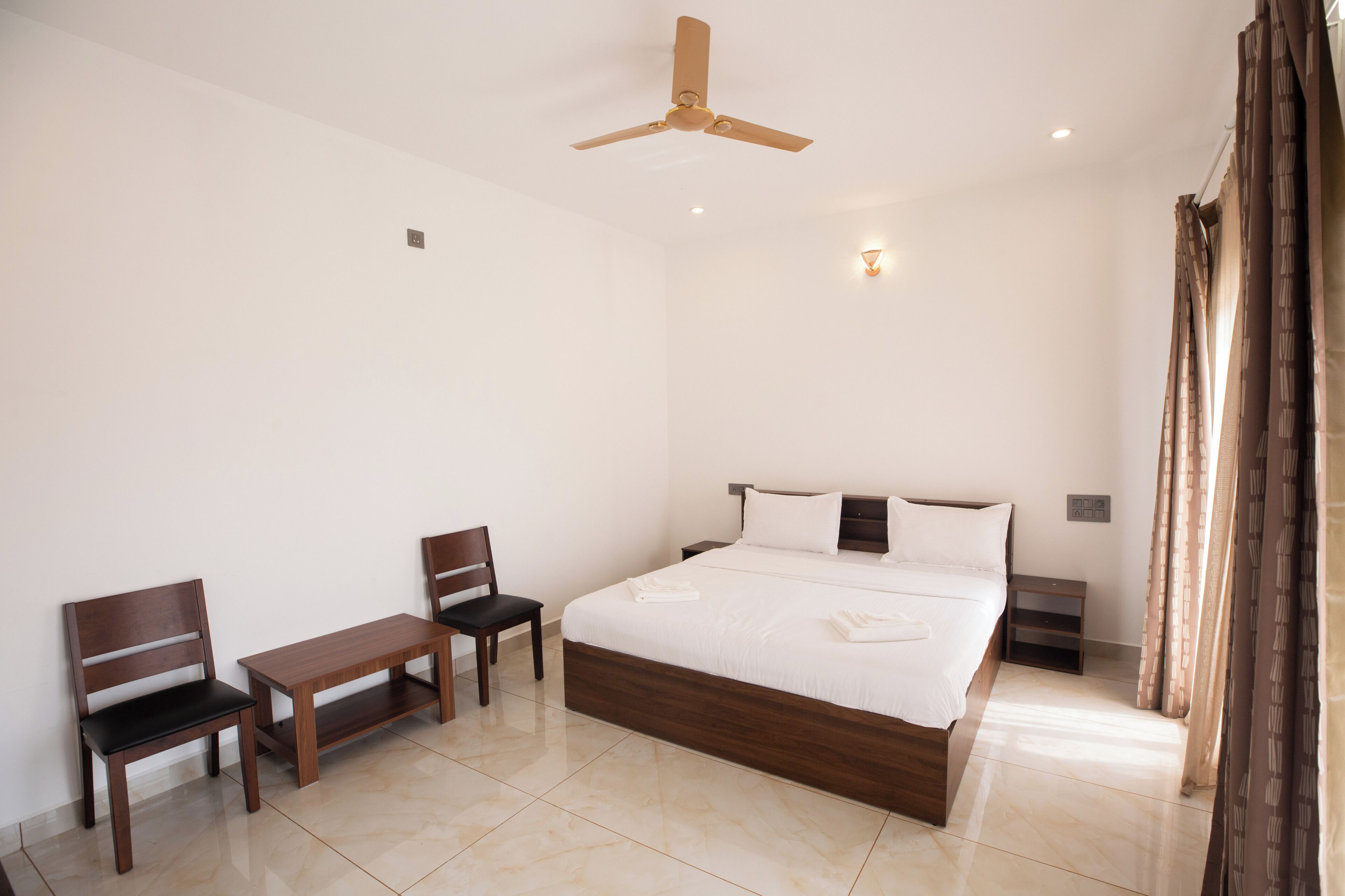 A hotel room with a bed, a desk, two chairs, a ceiling fan, and a nightstand.