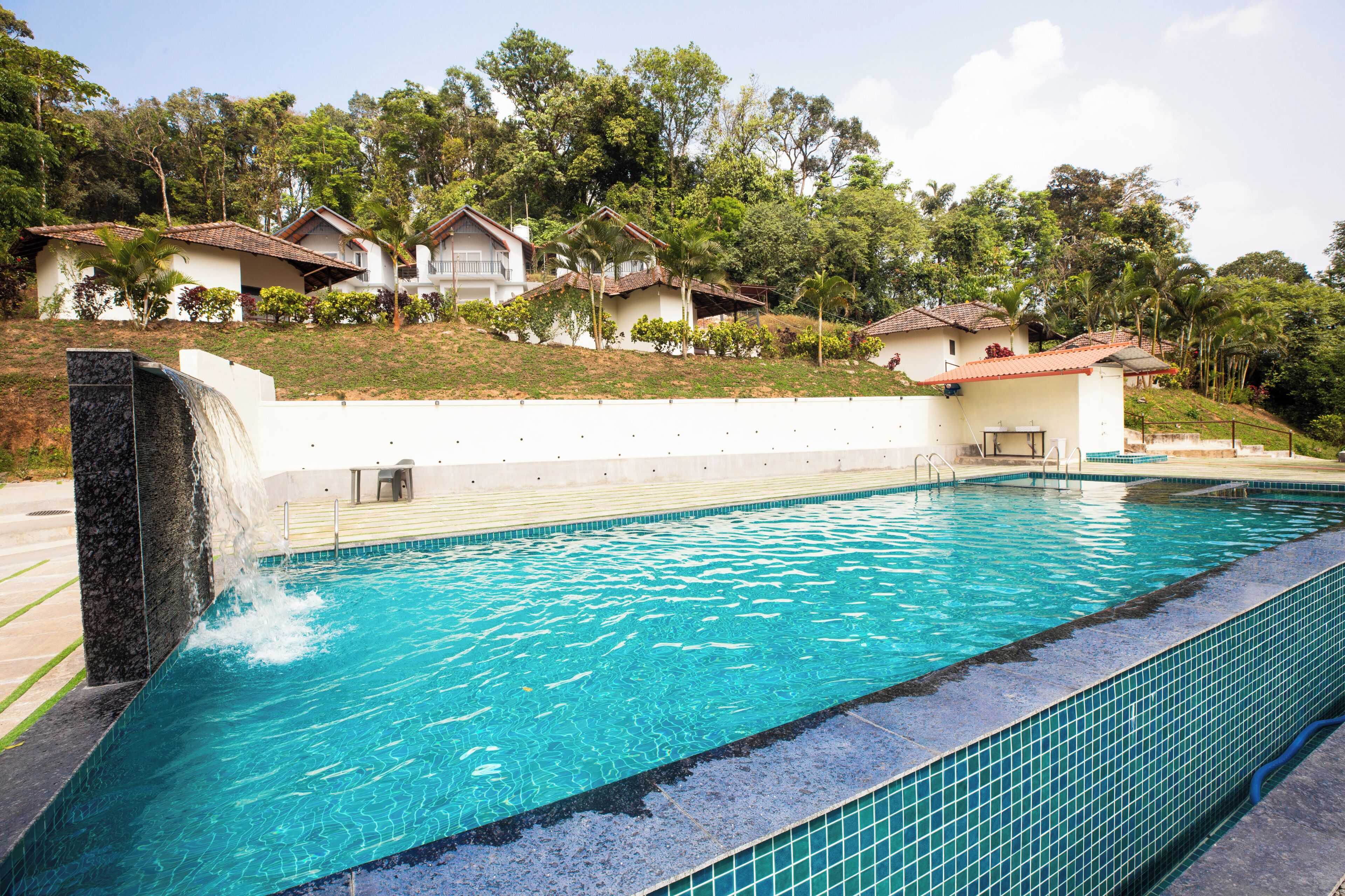 A swimming pool with a waterfall feature, surrounded by a white wall and a landscaped area with trees and a small building.