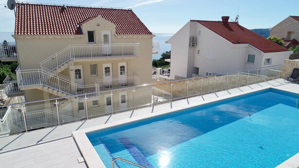 Una piscina con ringhiera in vetro, edifici vicini con tetti in tegole rosse e cielo sereno.