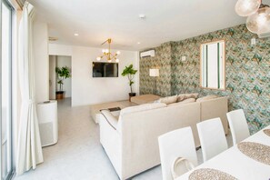 A modern living room with a sofa, dining table, and chairs. There is a TV mounted on the wall, a ceiling light fixture, and an air conditioning unit.