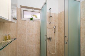 Ein Badezimmer mit Duschkabine aus Glas, einer Handbrause, einer weiteren Handbrause, einer Sitzgelegenheit aus Glas in der Dusche und einem Fenster mit einer Orchideenpflanze.