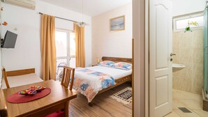 Un dormitori amb un llit, un escriptori amb una cadira, una finestra amb cortines, un quadre a la paret i un bany amb un lavabo.