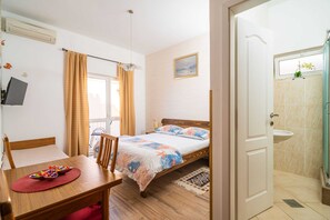 Ein Schlafzimmer mit Bett, Schreibtisch mit Stuhl, Fenster mit Vorhängen, einem Bild an der Wand und einem Badezimmer mit Waschbecken.