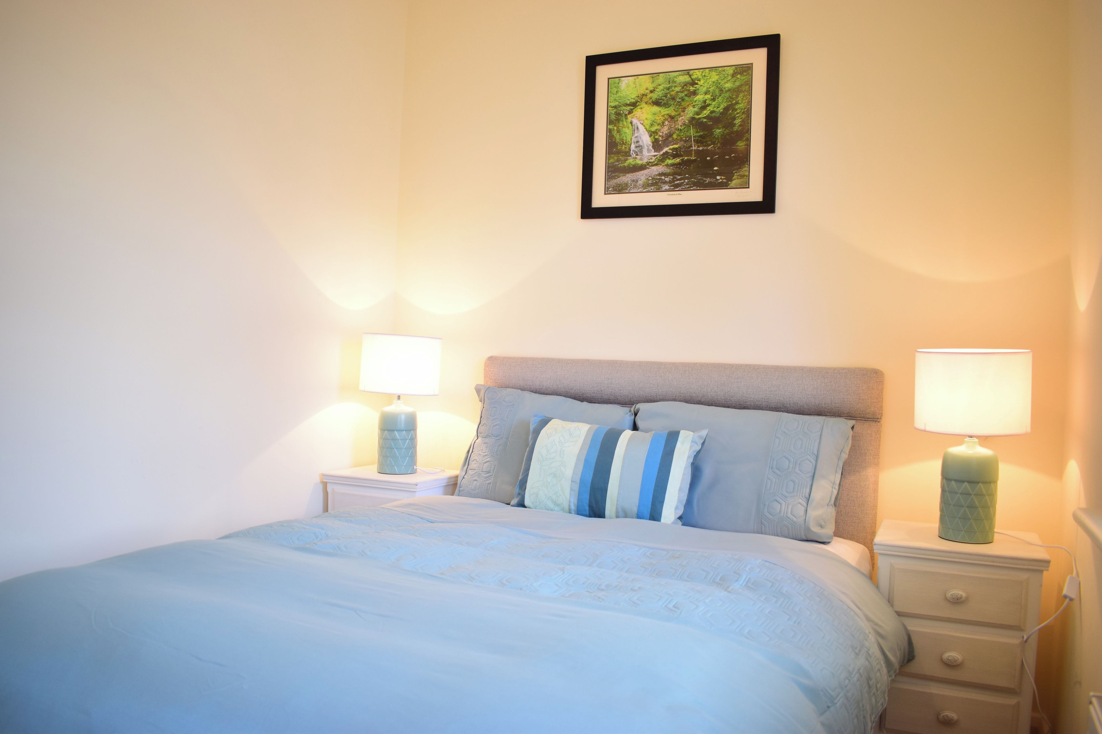 A bedroom with a bed, two bedside tables with lamps, a nightstand, and a framed picture on the wall.
