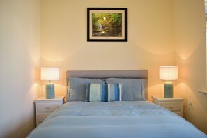 Un lit bien fait, recouvert d’une couvre-lit à motifs, avec deux lampes de chevet et un tableau encadré représentant un paysage naturel accroché au mur.