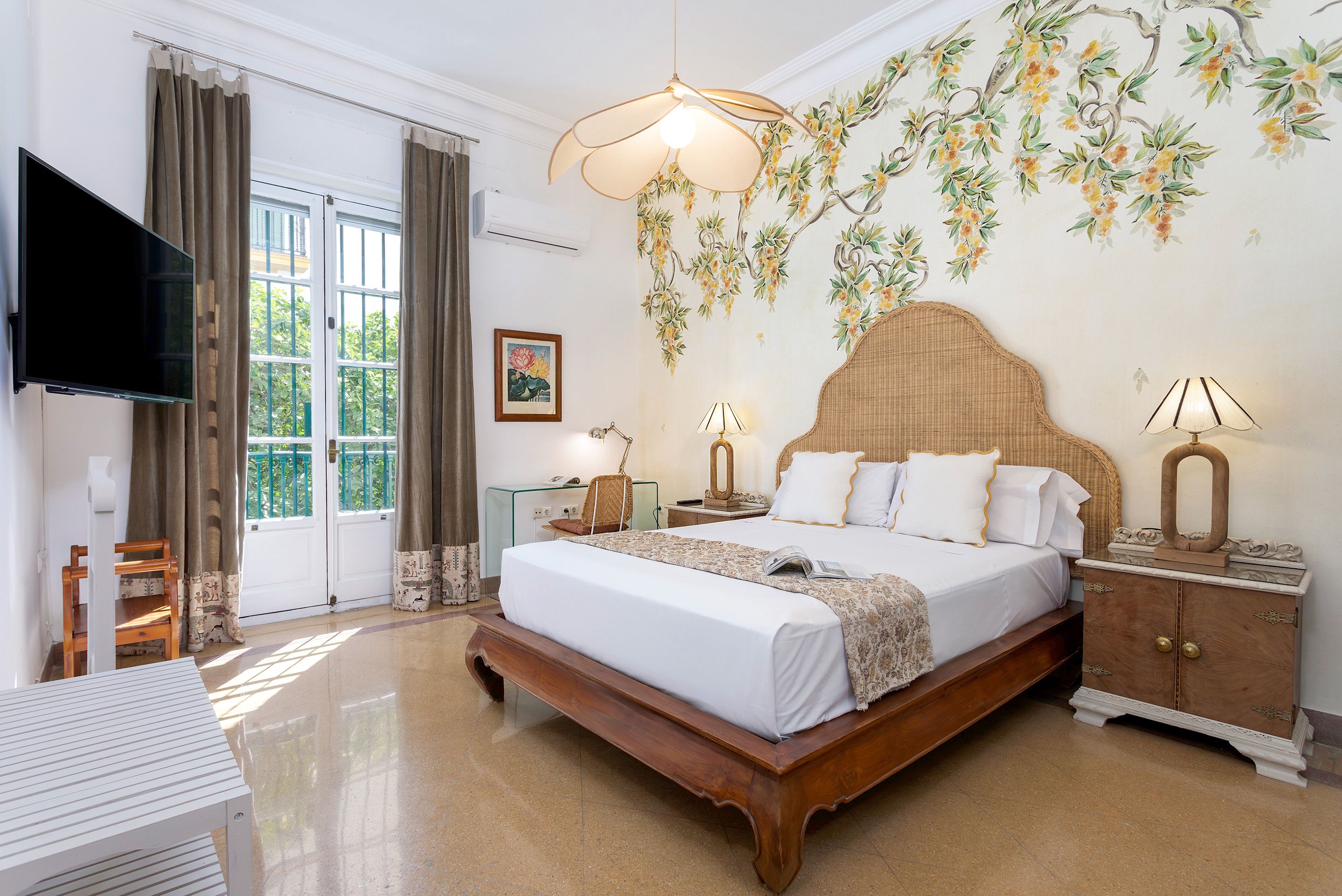 A hotel room with a large bed, a television, a window with curtains, a wall-mounted lamp, and a decorative wall hanging.