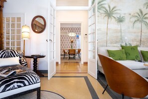 Un salón con un sofá estampado con diseño de cebra, una silla de madera, una mesa con una lámpara y un cuadro con palmeras.