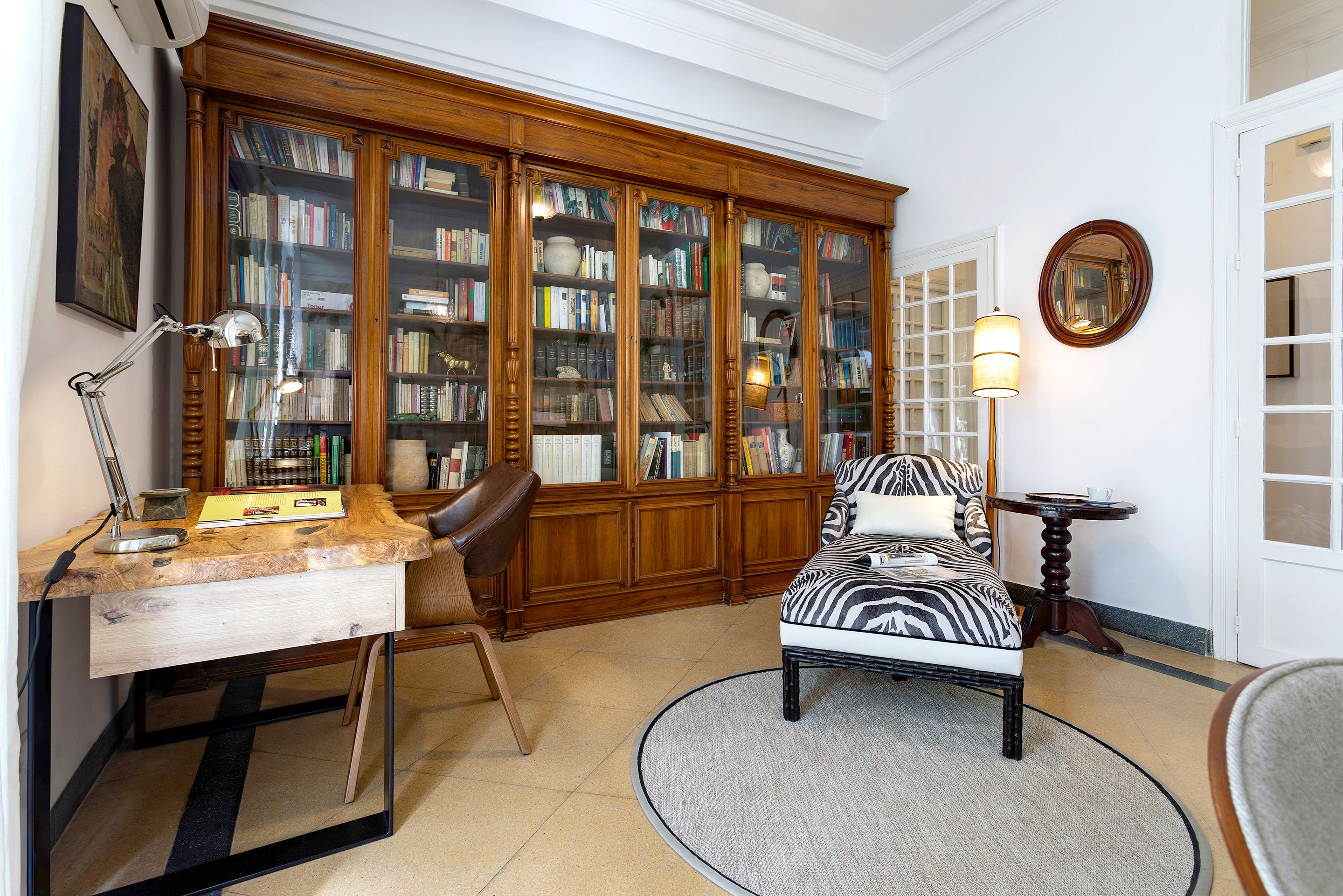 A room with a wooden bookcase, a desk with a lamp, a zebra-patterned chair, and a round table with a lamp.
