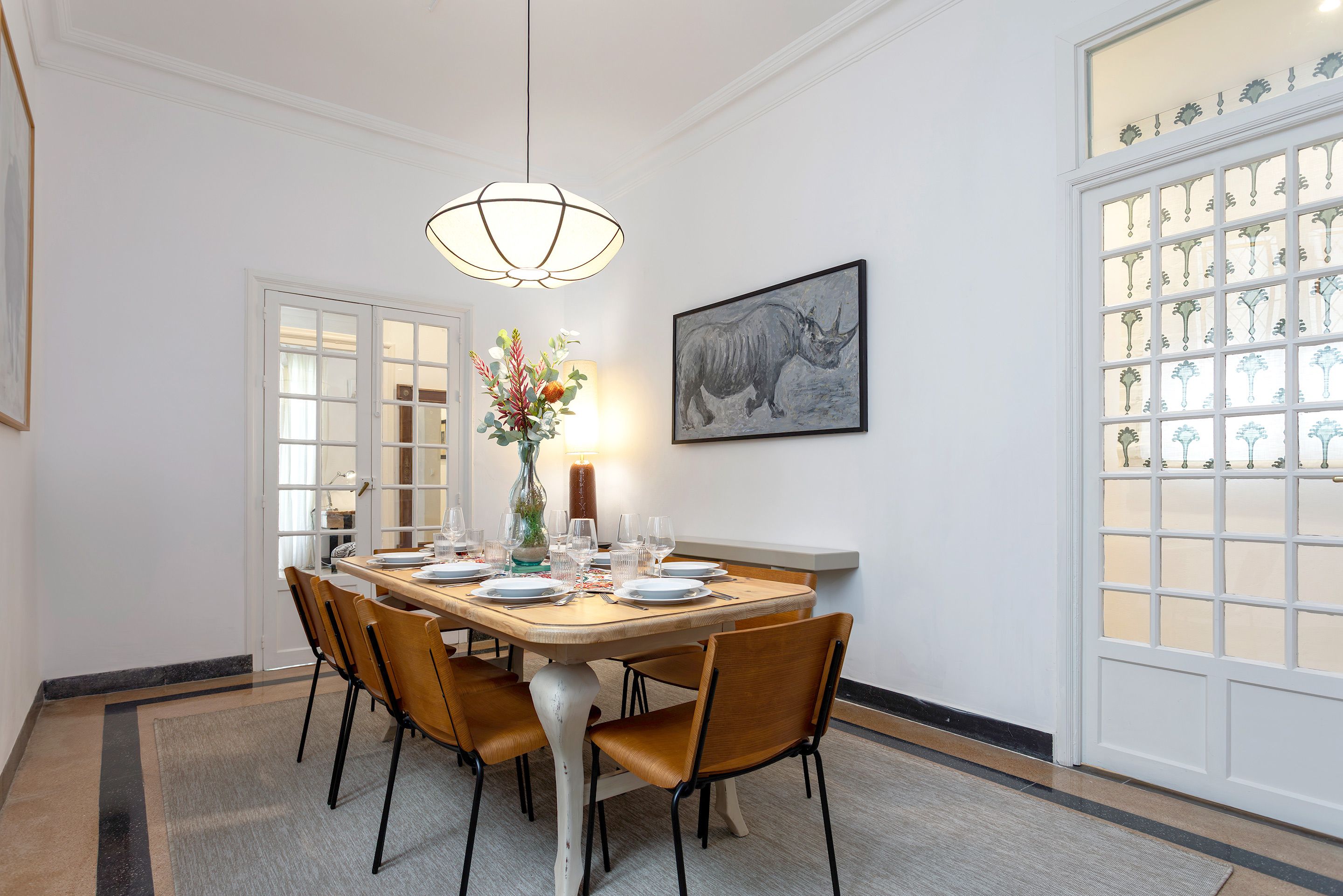 Une salle à manger avec une table en bois mise pour quatre personnes, entourée de chaises, un vase rempli de fleurs et un tableau accroché au mur.