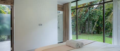 Un dormitorio moderno con una cama grande, ducha y vistas a un jardín exuberante a través de ventanales de gran altura.