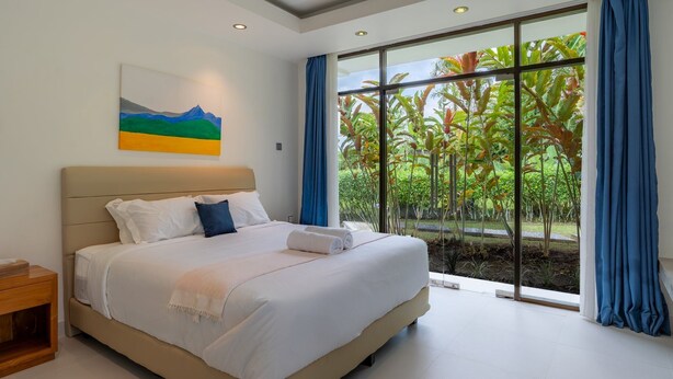 Une chambre moderne avec un grand lit, une table de chevet, un tableau et une vue sur la verdure grâce à de grandes fenêtres.