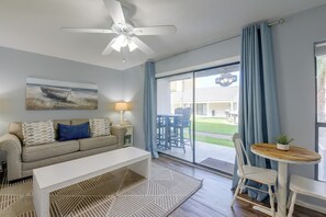 A living room with a sofa, a coffee table, a ceiling fan, a painting, a lamp, and a view of the outside. - Walk to Shore! PCB Condo w/ Pool & Hot Tub Access (Panama City Beach)