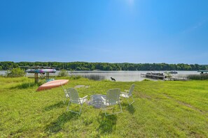 A grassy area with a row of white chairs and a canoe, overlooking a calm lake with boats and a dock. - Boating, Bonfires & Lake Views! Michigan Getaway (Ellsworth)