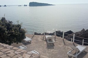 Eine Seeterrasse mit Steintreppe zu weißen Liegestühlen, mit Blick auf ruhiges Wasser und entfernte Felsenformationen.