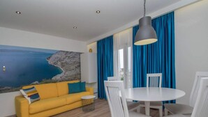 A room with a yellow sofa, white chairs, a round table, and a large window with blue curtains. - Apartments Antonio - Comfort One-bedroom Apartment With Balcony and Sea View - 4 (Tucepi)